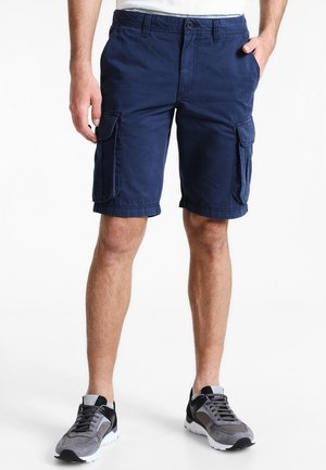 Mand iført marineblå cargoshorts og grå sorte sneakers stående med hænderne i lommerne på hvid baggrund.
