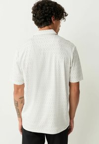 Balta trumpų rankovių marškiniai su šviesiai pilkų šešiakampių geometriniu raštu. Laisvas kirpimas su mažais apykakle ir lygia tekstūra.