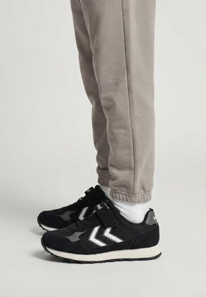 Ben iført beige joggerbukser og hvide sokker med sorte sneakers med hvide chevron-striber og velcrolukninger.