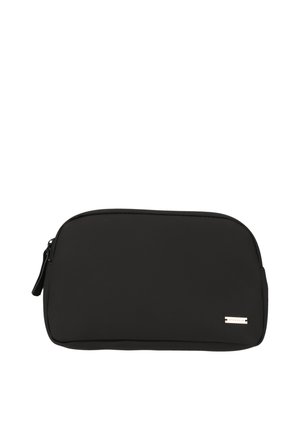 Femme aux cheveux bouclés portant un sweat beige et un pantalon, tenant une pochette noire sur un fond clair uni.