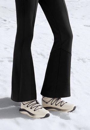 Bež pohodniški čevlji s črnimi vezalkami in teksturiranim podplatom, združeni s črnimi razširjenimi hlačami, na zasneženem ozadju.