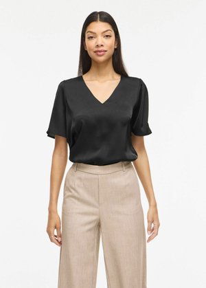 Kvinne med langt mørkt hår iført en svart V-hals kortermet bluse og beige bukser med høy midje og vide ben, stående mot en hvit bakgrunn.