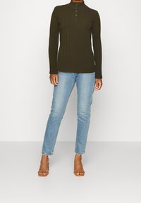 Dunkelgrünes, geripptes Langarmoberteil mit hohem Kragen und vier vorderen Knöpfen, kombiniert mit hellblauen Slim-Jeans und braunen, geschnürten Sandalen mit Absatz.