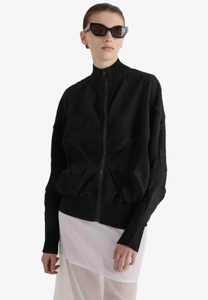 Kvinde iført sort tekstureret lynlåstrøje, hvid nederdel med gennemsigtigt lag og sorte solbriller, stående mod en ensfarvet baggrund.