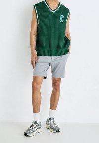 Zelený žebrovaný svetr bez rukávů s bílým lemem a písmenem 'C', doplněný o šedé šortky a šedé vzorované tenisky. Bílé kotníkové ponožky součástí.