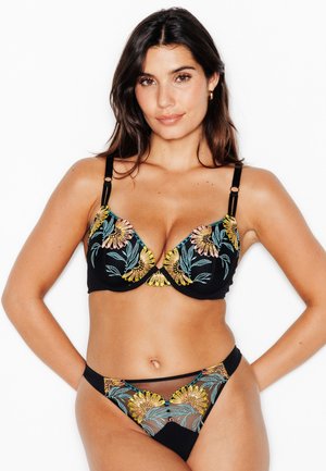 Femme portant un ensemble coordonné de soutien-gorge et culotte noirs avec une broderie florale colorée, debout sur un fond blanc uni.