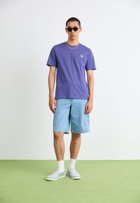 T-shirt violet, short en jean et chaussures à enfiler à carreaux. Lunettes de soleil et collier complètent la tenue décontractée. Fond texturé.