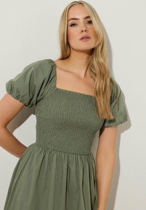 Olivengrünes Kleid mit kurzen, gepufften Ärmeln und einem quadratischen Ausschnitt. Texturierter geraffter Oberteil geht in einen fließenden Rock über. Weicher Baumwollstoff.