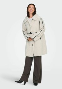 Cappotto strutturato di colore crema, con colletto, linee di accentuazione nere e chiusura a bottoni, abbinato a pantaloni scuri e stivali con tacco alto.