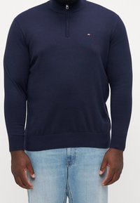 Homme portant un pull bleu marine à col zippé et un jean bleu clair, debout les bras détendus le long du corps devant un fond uni.