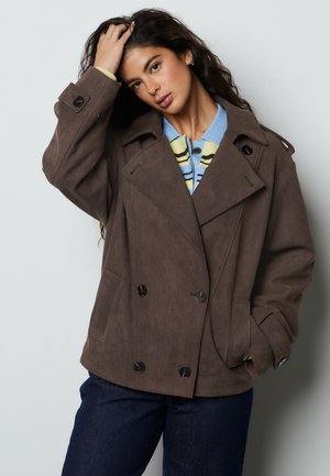 Nő hosszú, göndör hajjal, barna kétsoros kabátot visel, kék és sárga csíkos inget és sötét nadrágot, egyik kezét a fején tartva pózol.