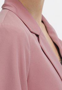 Pehmeän vaaleanpunainen bleiseri, jossa on klassinen kaulus, sileä pinta ja räätälöity istuvuus. Mallissa on puhtaat linjat ja hillityt saumausyksityiskohdat.