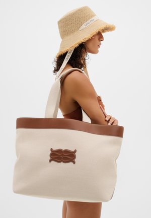 Naine pruunis ujumistrikoodis, kandes beežikat laiade äärtega mütsi, millel on tekst "MALINA", ning kandmas suurt beeži ja pruuni, nahkdetailidega ja logoga kandekotti.