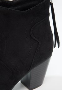 Gros plan sur une bottine en daim noir avec couture décorative et talon empilé épais sur fond blanc.