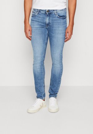 Mand iført lyseblå slim fit-jeans og hvide sneakers, stående mod en ensfarvet hvid baggrund.