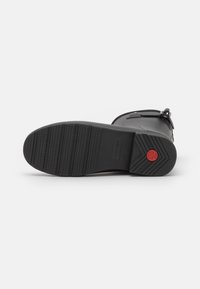 Suela de bota de goma negra con un patrón de relieve texturizado, un acento rojo en el talón y un detalle de correa cerca del tobillo.