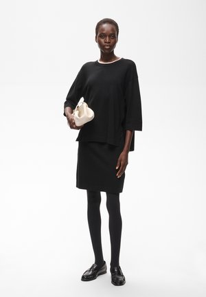 Femme en robe noire, collants et chaussures tenant un petit sac à main blanc sur un fond blanc uni.