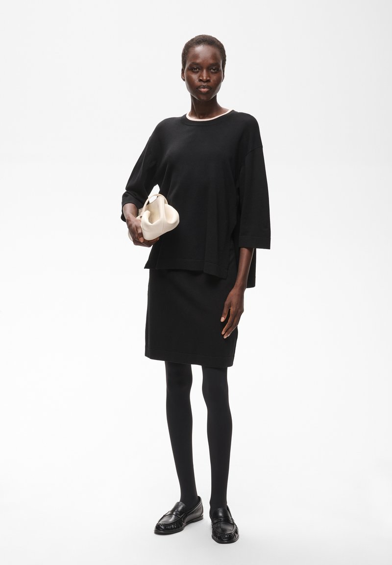 Femme en robe noire, collants et chaussures tenant un petit sac à main blanc sur un fond blanc uni.