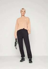 Mlada osoba s kratkom plavom kosom nosi bež majicu dugih rukava, crne trenirke, crne tenisice, drži crni konopac za preskakanje na jednostavnoj pozadini.