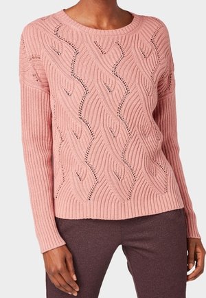 Femme portant un pull en maille rose à manches longues avec un motif texturé ondulé, associé à un pantalon marron foncé.