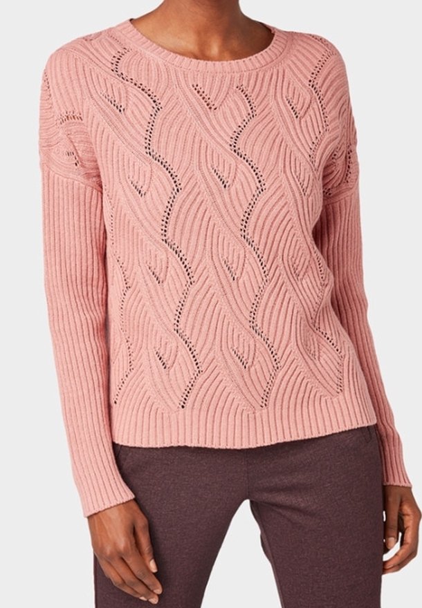 Femme portant un pull en maille rose à manches longues avec un motif texturé ondulé, associé à un pantalon marron foncé.
