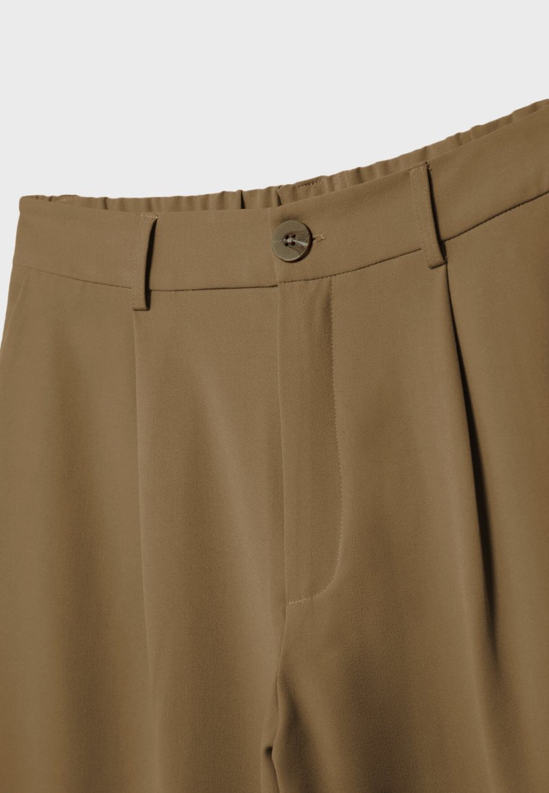 Gros plan sur un pantalon marron avec une fermeture à bouton, des passants de ceinture et un détail de taille élastique sur un fond uni.