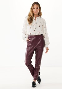 Vrouw in een witte blouse met lange mouwen en kleine bloemenprints, bordeaux leren broek en zwarte instappers, staand met één hand in de zak.