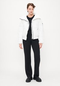 Witte pufferjas met hoge kraag en ritssluiting, gedragen over een zwarte top en gecombineerd met zwarte wijde broek en plateau schoenen.