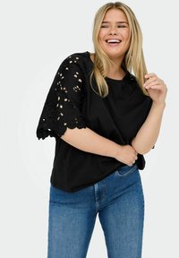 Black top with short, lace sleeves featuring floral cutouts. Paired with blue denim jeans. Smooth fabric, casual design.