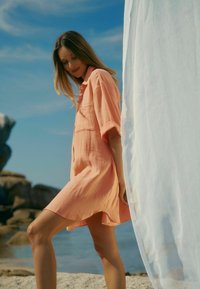 Pfirsichfarbiges, leichtes Leinenhemdkleid mit kurzen Ärmeln, einer Brusttasche und lockerer Passform, vor einer Strandkulisse.