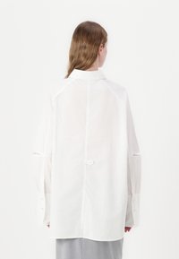 White cotton shirt with a point collar, long sleeves, and cut-out detail at the cuffs. Smooth texture, straight cut, simplicity in design.