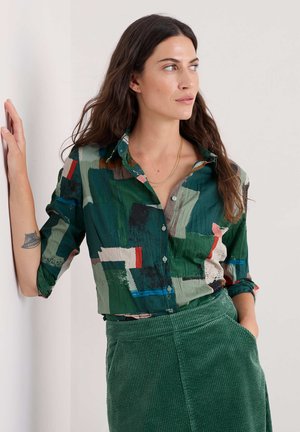 Vrouw met lang haar leunend tegen een muur, gekleed in een button-up shirt met een multicolored abstracte print en een groene corduroy rok.