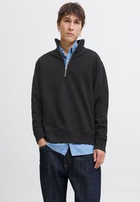 Noor mees lühikeste pruunide juustega, kannab rõngaskõrvarõngaid, musta poole luku jahekat sinise särgi kohal ning tumedaid teksapükse, seisab ühe käega taskus.