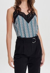 Ärmlös topp med en svart spetsdetalj, som har ett geometriskt mönster i blått och vitt, ihop med högmidjade svarta byxor med ett bälte.