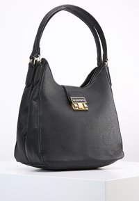 Svart läderhandväska med texturerad yta, detaljer i guld, och ett handtag på toppen. Har en klaffstängning med en logodetalj.