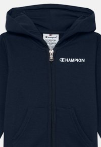 Jūras zila kapuce ar priekšējo rāvējslēdzēju, divām kabatām un baltu "CHAMPION" logotipu. Izgatavota no mīksta, gluda auduma, piemērota zīdaiņiem.