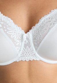 Vitt lingeribehå med blommönstrad spetskant, en slät koppdesign och en liten rosettaccent framtill. Mjuk textur.