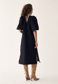 Femeie purtând o rochie midi texturată neagră cu mâneci bufante, detaliu cu panglică la spate, slit lateral și cizme maro deschis cu toc până la genunchi, stând cu fața spre spate.