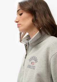 Sudadera de color gris claro con cremallera y cuello alto, que presenta el texto azul "SUPERDRY ATHLETICS" y un acento de bandera estadounidense. Forro interior a rayas.