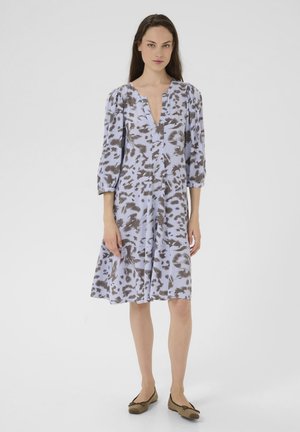 Robe bleu clair avec des motifs abstraits marron, en V, manches trois-quarts, et une silhouette évasée, portée avec des chaussures plates marron.