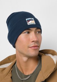 Gorro de punto azul marino con un parche de logo tejido que presenta un diseño paisajístico. Textura suave, ajuste ceñido y patrón acanalado en todo el gorro.