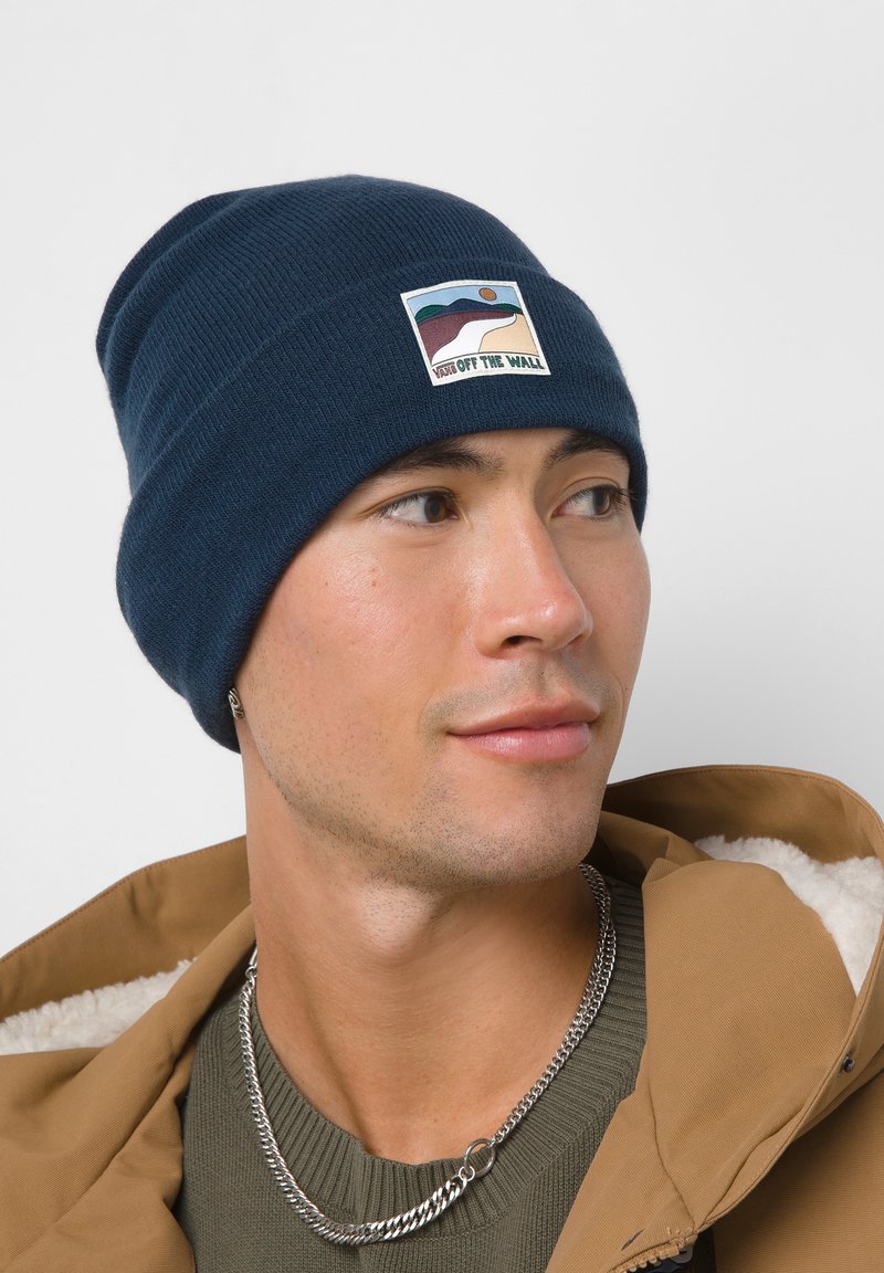 Gorro de punto azul marino con un parche de logo tejido que presenta un diseño paisajístico. Textura suave, ajuste ceñido y patrón acanalado en todo el gorro.