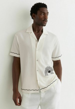 Mann trägt ein weißes Kurzarmhemd mit schwarzen wellenförmigen Akzenten und abstraktem schwarzem Muster am Saum, schaut nach rechts.