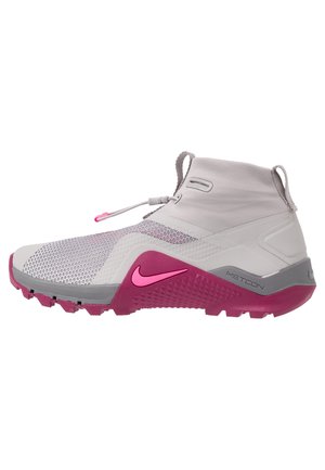 Detail šedé a růžové atletické boty Nike Metcon s prodyšným síťovaným svrškem, vysokým límcem kolem kotníku a texturovanou podrážkou pro lepší přilnavost.