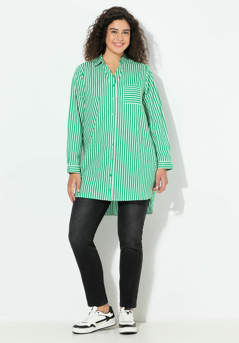 Camisa a rayas verdes y blancas con un bolsillo en el pecho, cierre de botones en la parte delantera, mangas largas y dobladillo curvado, combinada con pantalones negros y zapatillas.