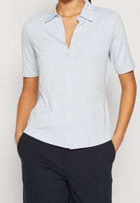 Donna che indossa una camicia azzurra chiara a maniche corte, infilata in pantaloni blu scuro, con le mani in tasca, su uno sfondo neutro.