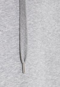 Close-up van een grijze hoodie-touw met een geweven textuur en een zilveren metalen aglet aan het einde, tegen een grijze stof.