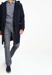 Cappotto lungo in misto lana blu navy con chiusure a toggles, indossato sopra una camicia a maniche lunghe azzurra con fantasia, abbinato a pantaloni grigi e scarpe blu.