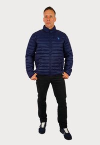 Marineblå pufferjakke med lynlås foran, lodret quiltet design, sidelommer og et logo på brystet, kombineret med sorte jeans og blå sneakers.