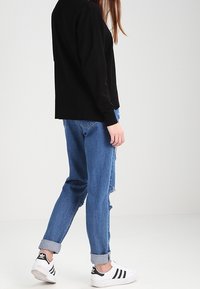 Zwarte oversized trui, blauwe distressed jeans opgerold bij de boorden, witte sneakers met zwarte strepen, casual outfit zijdelings bekeken.